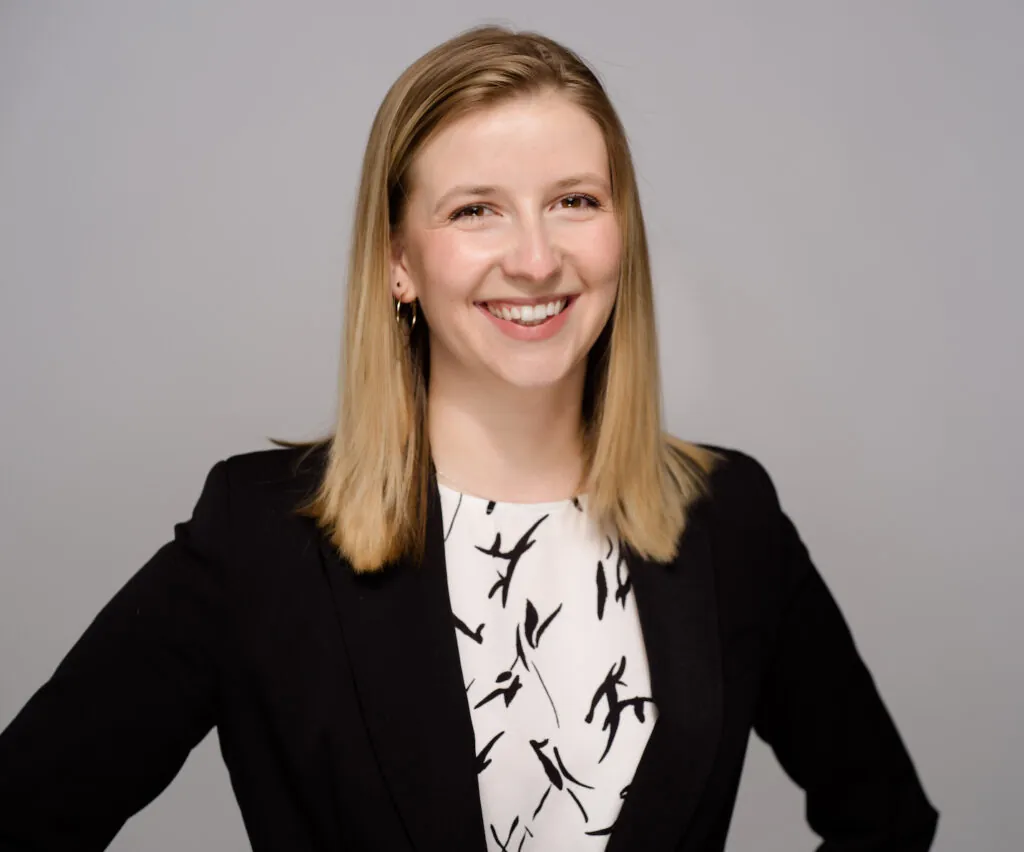 A professional headshot of an unnamed person, smiling, with short hair, wearing a dark suit.