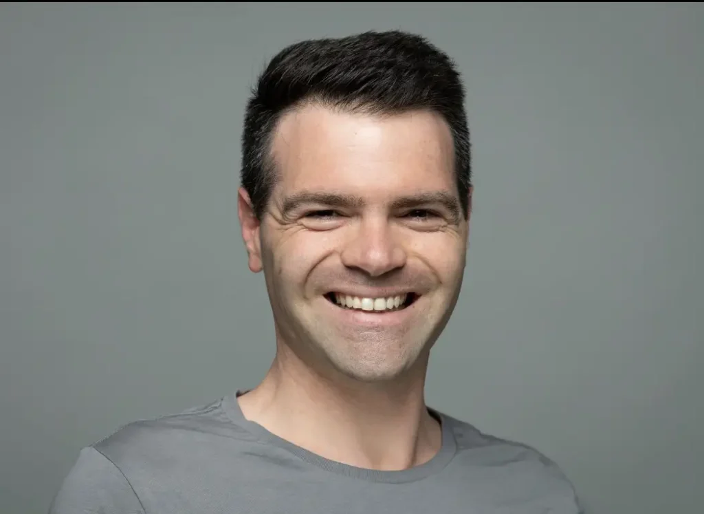 Casual photo of a person standing outside, smiling, with short hair, wearing a blue jacket and a white shirt, against a blurred background.