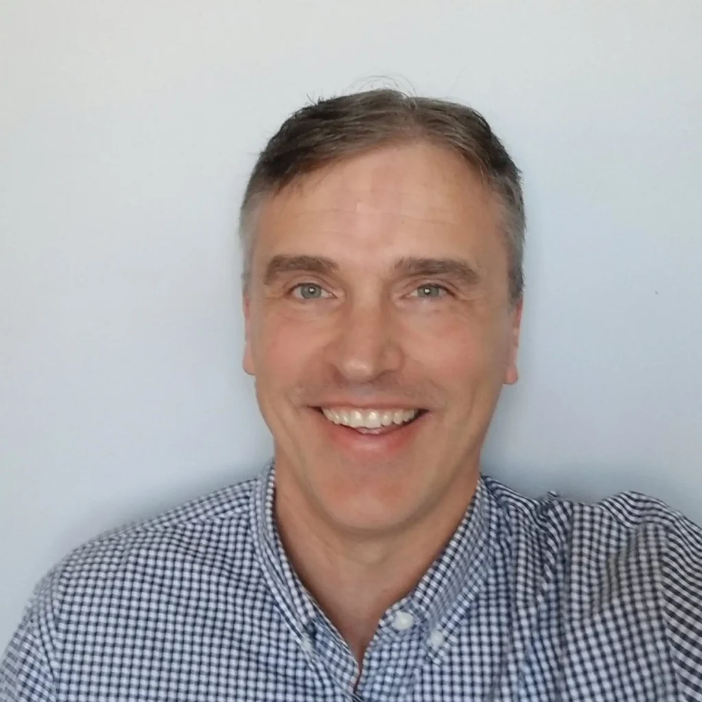 Professional headshot of a person with short hair, smiling, wearing a dark blazer and a light shirt, against a plain background.