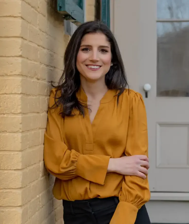 Professional headshot of a person, smiling, with short hair, wearing a dark blazer and a light shirt, against a plain background.