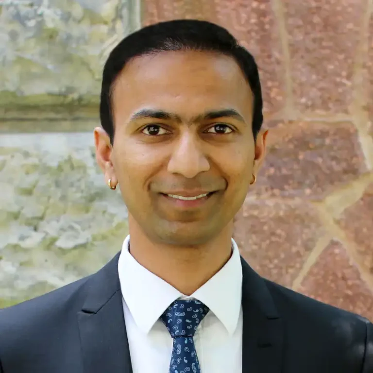 Professional headshot of Ashwin, smiling, with short hair, wearing a dark blazer and a light shirt, against a plain background.