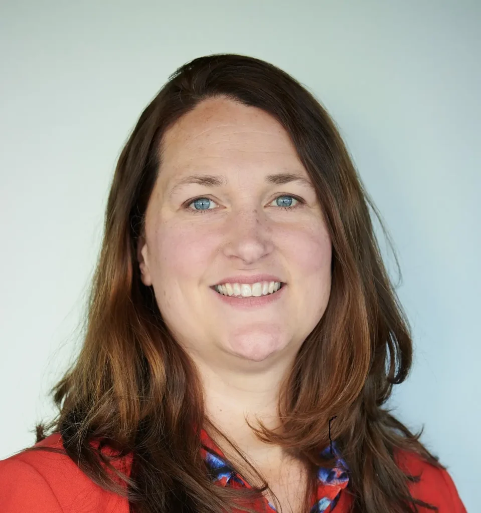 Professional headshot of Ilsa Webeck, smiling, with shoulder-length hair, wearing a dark blazer and a light shirt, against a plain background.