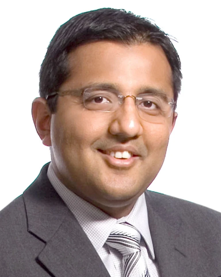 Professional headshot of Tejas Gandhi, smiling, wearing a blue suit jacket and a white shirt, with a blurred background.